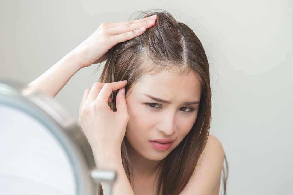 A young Asian woman with thinning hair looks in a mirror, concerned.